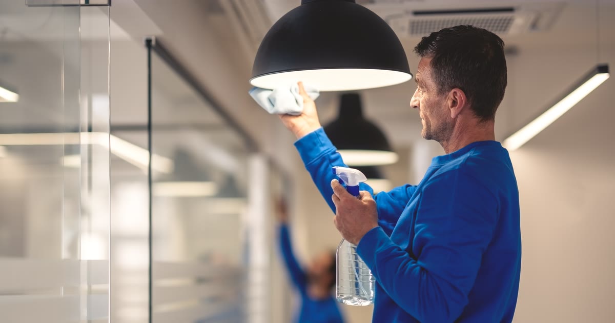 Reinigungskraft wischt eine Leuchte in einem Büro ab